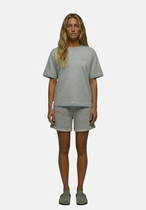 Femme debout, de face, portant un t-shirt oversize gris clair, un short assorti et des chaussures slip-on gris foncé sur un fond uni.