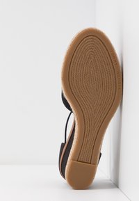 Semelle de chaussure style espadrille avec motif tissé, semelle en caoutchouc beige et dessus noir, positionnée contre un mur blanc d’angle.