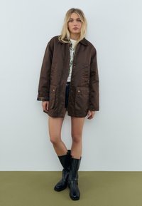 Ung kvinde med blondt hår iført en brun oversized jakke, en hvid t-shirt med grafik, denimshorts og sorte støvler til midt på læggen, der står op ad en væg.