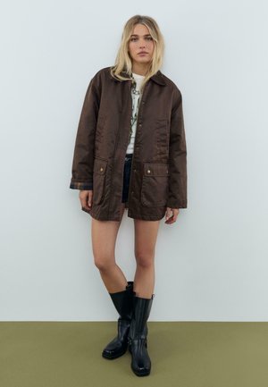 Młoda kobieta z blond włosami, ubrana w brązową, oversize'ową kurtkę, białą koszulkę z grafiką, dżinsowe szorty i czarne botki do połowy łydki, stojąca przy ścianie.