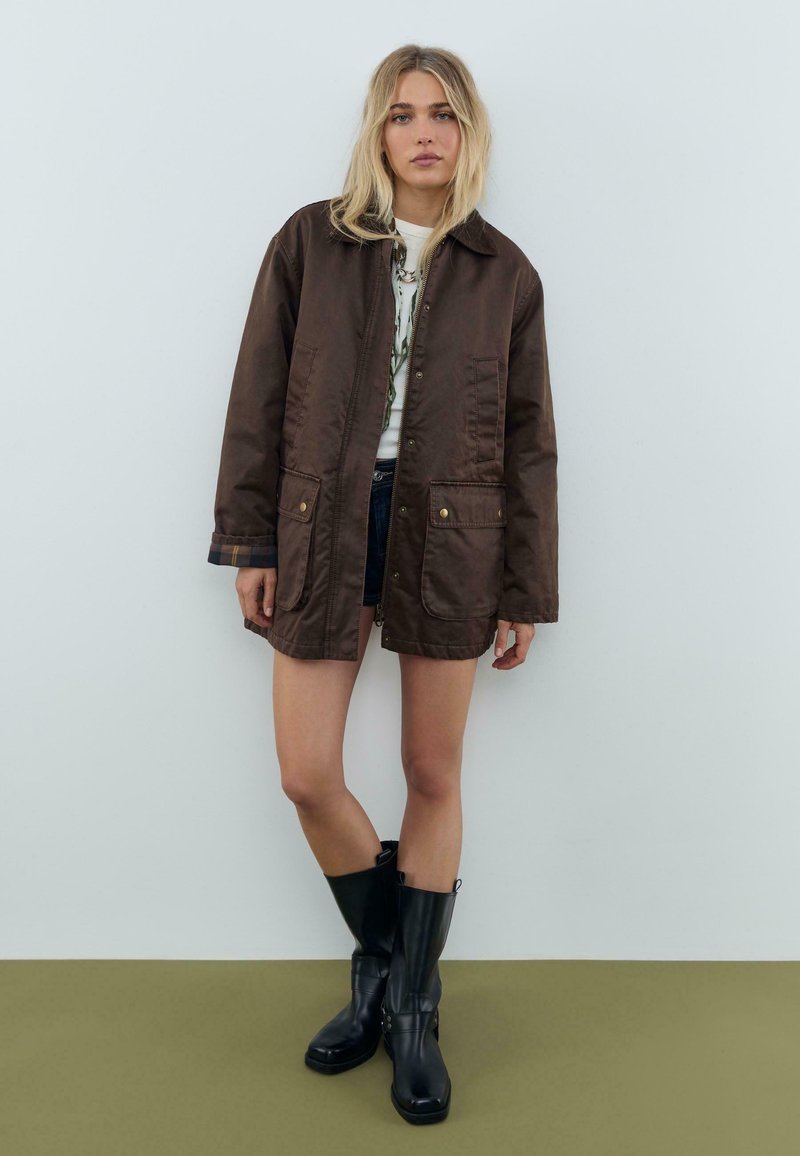 Ung kvinde med blondt hår iført en brun oversized jakke, en hvid t-shirt med grafik, denimshorts og sorte støvler til midt på læggen, der står op ad en væg.