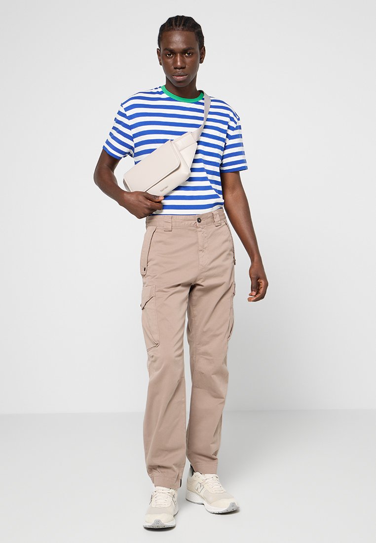 Gestreept blauw-wit T-shirt met groene accenten, beige cargobroek met zakken en een licht crossbodytas. Sneakers maken de look compleet.