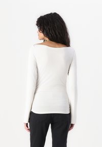 White long-sleeve top with a relaxed fit, round neck, and gathered detail at the waist, paired with black pants. Smooth texture.