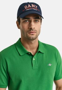 Groene polo met kraag en knopen, voorzien van een klein GANT-logo. Vergezeld van een marineblauwe pet met rode geborduurde tekst.
