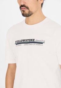 Mies, joka käyttää valkoista T-paitaa, jossa lukee "Yellowstone National Park Wyoming Yhdysvallat" etupuolella, valkoista taustaa vasten.
