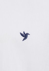 Valge kangas, millel on väike tumesinise värvusega tikandiga kolibri disain. Kangas tundub sile ja kerge.