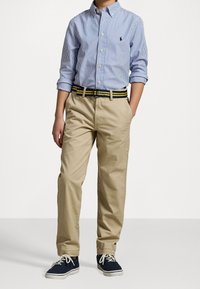Garçon portant une chemise à boutons bleu et blanc rayée rentrée dans un pantalon beige avec une ceinture rayée noire et jaune et des baskets bleu marine.