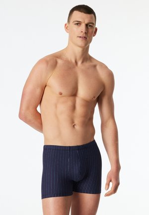 Muscular man standing with one hand behind back, wearing navy blue striped boxer briefs against plain white background.