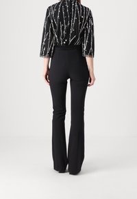 Personne portant un pantalon noir à taille haute et évasé ainsi qu'un haut noir transparent avec des rayures verticales en paillettes argentées, debout, vue de dos.
