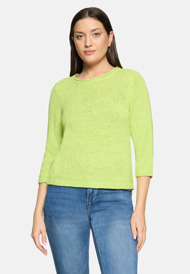 Femme aux cheveux raides et foncés portant un pull tricoté vert vif et un jean bleu, sur un fond clair uni.