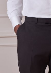 Pantaloni neri in tessuto strutturato con fronte piatto, tasche laterali e aspetto elegante, abbinati a una camicia bianca con bottoni.