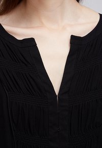 Czarna bluzka z dekoltem w kształcie V, marszczonymi detalami oraz teksturowanym przeszyciem z przodu. Materiał wydaje się miękki i lekki.