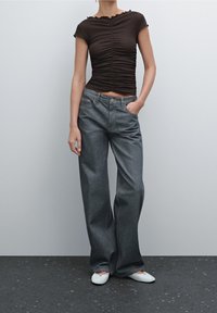 Brown fitted top with gathered details, paired with loose dark denim jeans and white flat shoes, set against a neutral background.