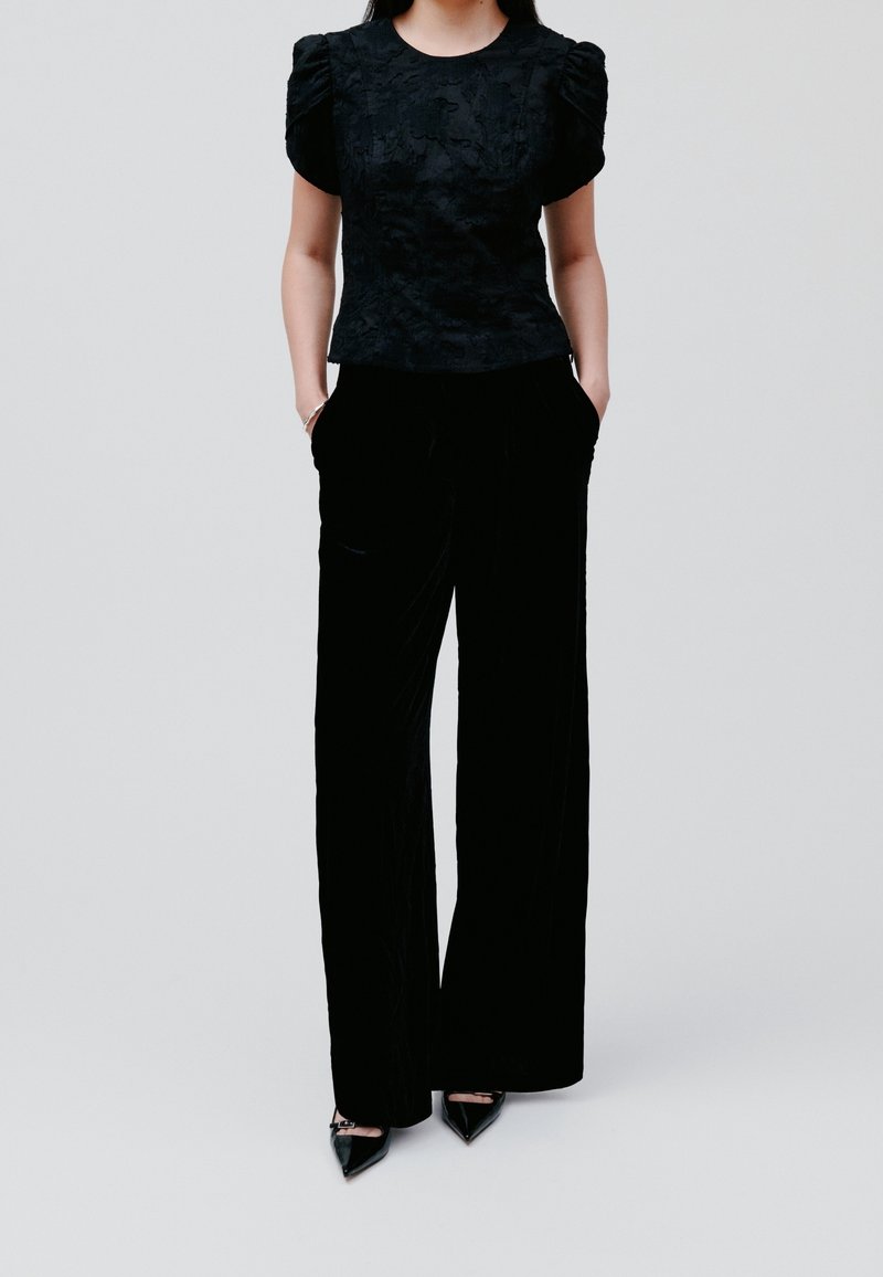Femme portant un haut noir texturé à manches bouffantes, un pantalon large noir et des talons pointus noirs, debout avec les mains dans les poches.