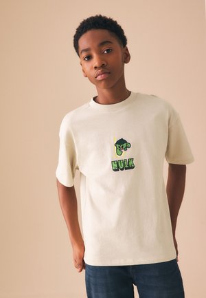 Fiatal fiú rövid, göndör hajjal, bézs pólót visel zöld Hulk grafikával és sötétkék farmerral, egyszínű háttér előtt áll.