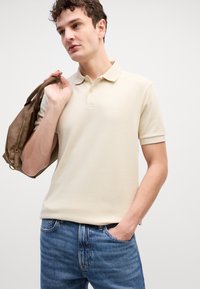 Beige rövid ujjú pólóing gallérral, a mellkason diszkrét "BOSS" logó hímezve. Kék farmernadrággal párosítva.