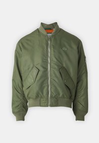 Jachetă bomber de culoare verde măslin, cu fermoar pe partea din față, manșete ribbed și două buzunare frontale. Prezintă un logo mic pe pieptul stâng și o captuseală interioară în contrast.
