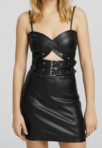 Žena v černých mini šatech z umělé kůže s křížovým dekoltem, výřezem v pase, tenkými ramínky a detailem dvojité přezky na pásku.