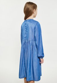 Denimkjole i lyseblå med lange puffærmer, en tætsiddende overdel og en samlet nederdel. Bagsiden har en række af stofbeklædte knapper.