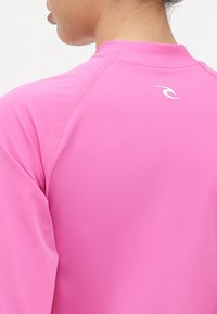 Ružičasta majica dugih rukava s visokim ovratnikom i bijelim logotipom na leđima. Glatka tekstura i dizajn rukava raglan za poboljšanu pokretljivost.