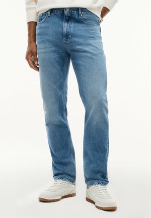 Mand iført lyseblå jeans med lige ben, hvide sneakers og en hvid langærmet top, står med den ene hånd i lommen.