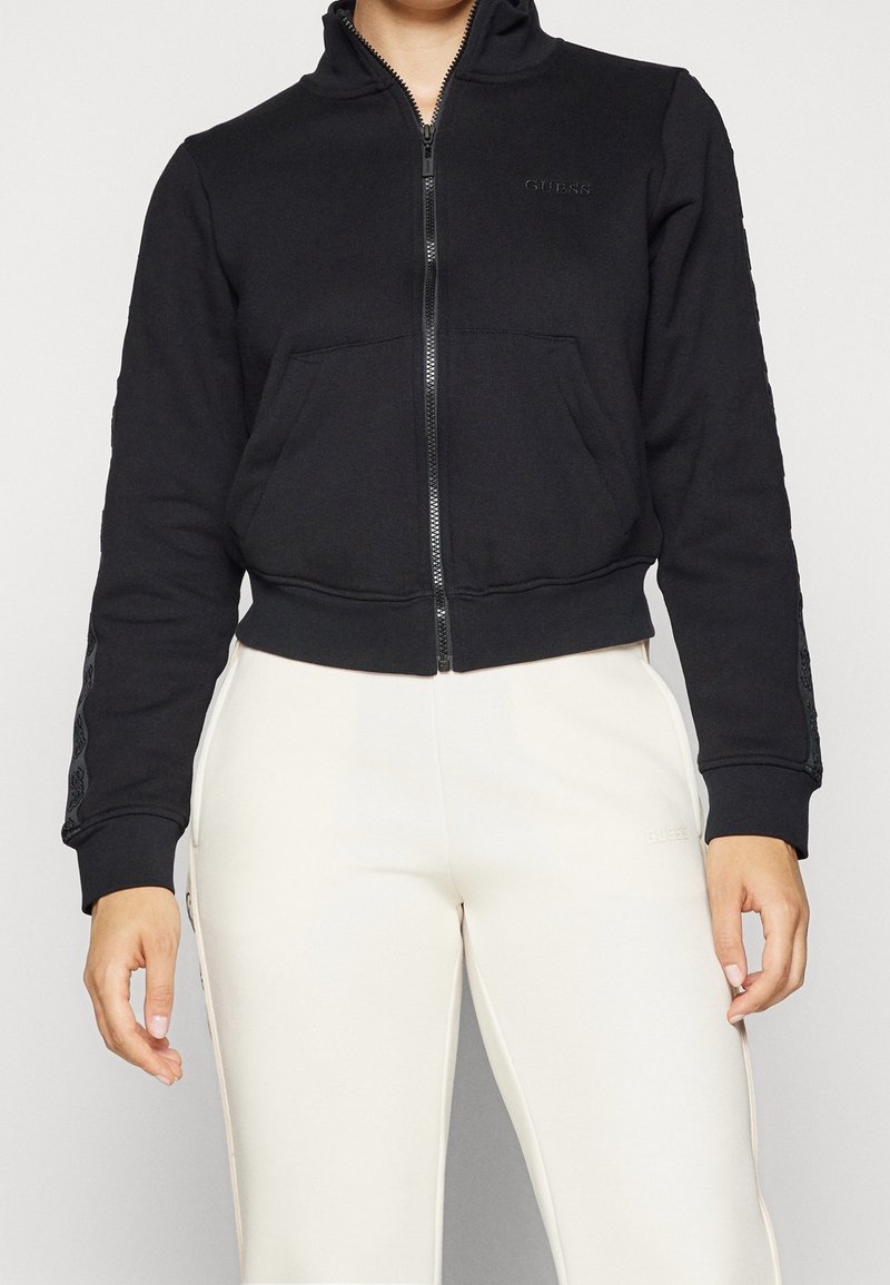 Personne portant un sweat à capuche noir zippé avec des poches avant et un pantalon blanc, debout devant un fond clair uni.