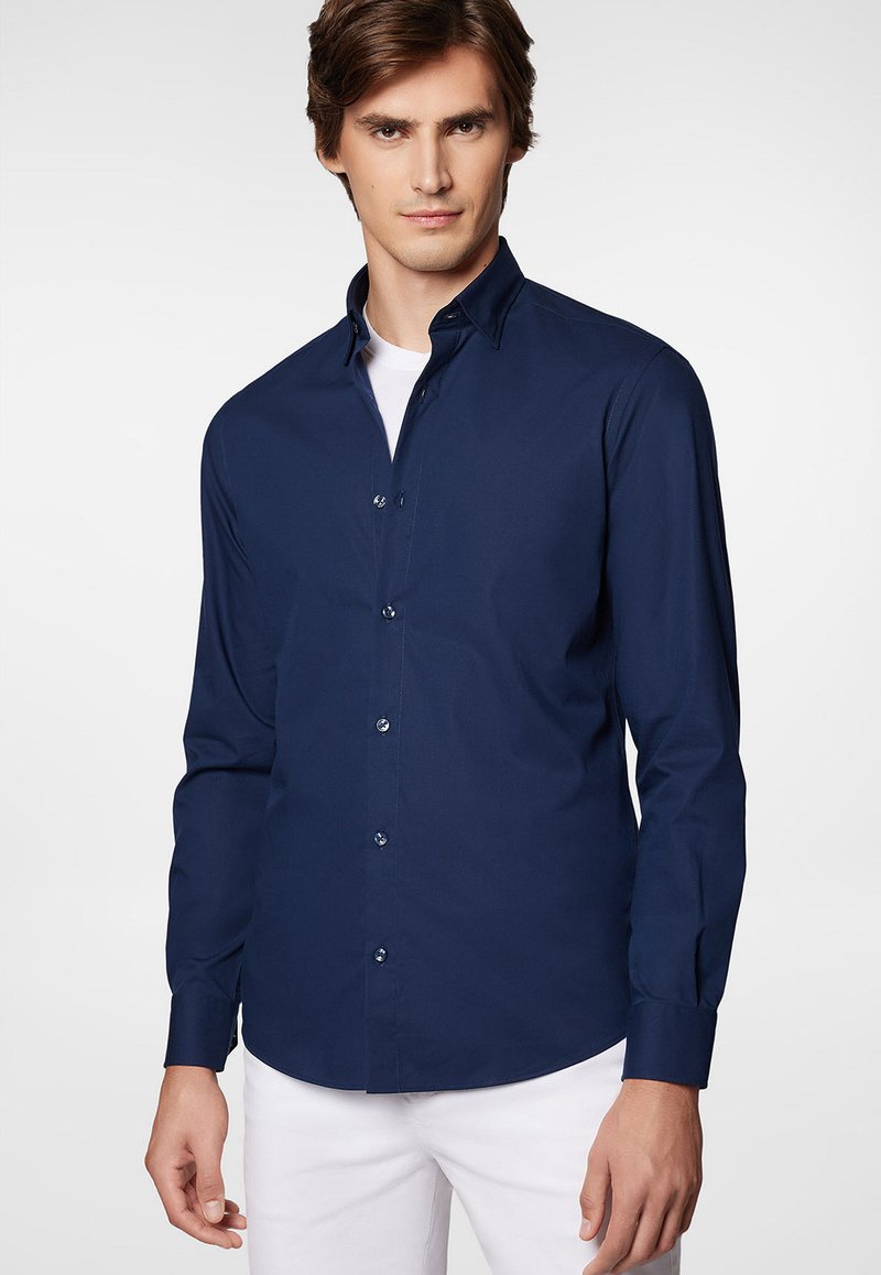 Granatowa koszula z długim rękawem i kołnierzykiem z guzikami, z przodu zapinana na guziki, o gładkiej teksturze materiału. Nosi się ją z białymi spodniami.