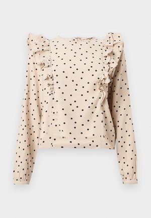 Blouse beige à manches longues avec des pois noirs, ornée de panneaux à volants descendant des épaules jusqu'à l'avant, et poignets boutonnés.