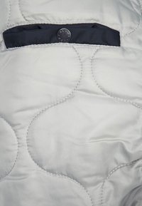 Tissu matelassé gris clair avec des coutures circulaires et une poche noire garnie d'un bouton gravé "ALPHA" sur le rabat.