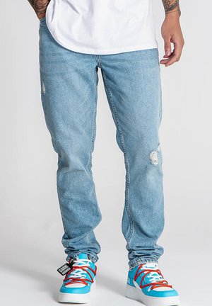 Jeans de mezclilla azul claro con corte ajustado, que presentan detalles desgastados y dobladillos deshilachados, combinados con zapatillas azul brillante y cordones rojos.