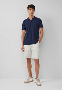 Marineblaues Poloshirt mit V-Ausschnitt, kombiniert mit hellbeigen strukturierten Shorts. Das Modell trägt weiße Sneakers mit beige- und marineblauen Akzenten.