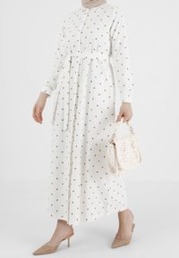 Robe blanche à manches longues avec des pois marron, devant à boutons, ceinture à la taille, et un sac à main en crème texturé tissé. Chaussures à talons beiges.