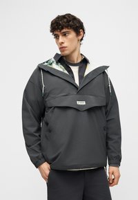 Νεαρός με σγουρά μαλλιά που φοράει σκούρο γκρι πουλόβερ Columbia με λουλουδένιο εσωτερικό κουκούλας, στέκεται μπροστά σε απλό φόντο.