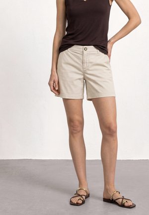 Kvinde iført beige knælange shorts, mørk ærmeløs top og brune sandaler, stående mod en hvid væg.