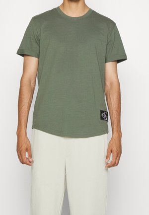 Mand iført en olivengrøn kortærmet t-shirt med Calvin Klein-logo på kanten og lysebeige bukser mod en ensfarvet baggrund.