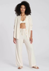 Chemise boutonnée beige clair sur un bralette assorti et un pantalon large. Texture douce, coupe décontractée et taille à cordon. Portée avec des sandales plates.