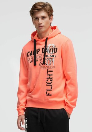 Junger Mann trägt einen leuchtend orangenen Kapuzenpullover mit schwarzem Text und Grafiken, kombiniert mit schwarzen Hosen, steht vor einem schlichten hellen Hintergrund.