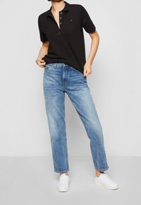 Schwarzes Poloshirt mit Knopfleiste und Logo, kombiniert mit hellblauen, gerade geschnittenen Jeans und weißen Sneakers. Einfaches, lässiges Outfit mit klaren Linien.