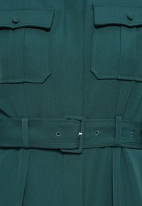 Tume roheline kangas, millel on kaks klapptaskut ja sobiv vöö ristkülikukujulise klambriga, mis rõhutab vöökohta.