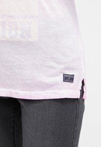 Ljusrosa bomulls t-shirt med en synlig logopatch i fållen, rundade kanter och sidofickor. Kombinerad med mörkgrå jeans.