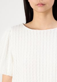 Hvit teksturert bluse med vertikale striper, med rund hals og pufferm. Stoffet ser lett og litt gjennomsiktig ut.
