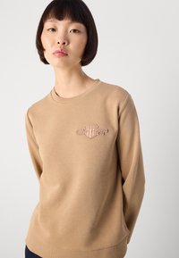 Giovane donna con capelli corti scuri che indossa una felpa beige con un piccolo emblema sul petto, in piedi davanti a uno sfondo chiaro e uniforme.