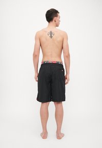 Black swim shorts with a decorative red accent on the side, featuring a white branded elastic waistband and a back pocket.