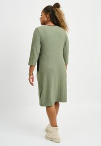 Grön knälång klänning med texturerad tyg, trekvartsärmar, sidofickor och svarta accenter. Matchad med ljusa ankelboots.