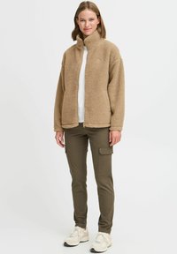 Veste en polaire beige avec col montant, fermeture éclair sur le devant, surface texturée. Associée à un pantalon cargo olive et des baskets légères. Palette de couleurs neutres.