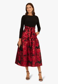 Schwarzes Langarmoberteil mit einem roten, floral gemusterten Rock. Der Rock hat eine Schleife an der Taille und besteht aus einem satinähnlichen Material.