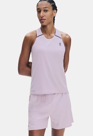 Frau mit geflochtenem Haar, die ein hellviolettes ärmelloses Sporttop und passende Shorts trägt, steht mit den Armen hinter dem Rücken.