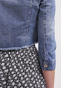 Žena v zkrácené modré džínové bundě s třepeným lemem a černé sukni s bílým květinovým vzorem, ruka uvolněně vedle těla.