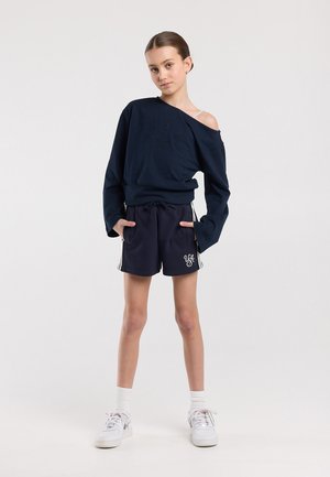 Meisje staand met handen in haar zakken, draag een marineblauwe off-shoulder trui, marineblauwe korte broek met witte strepen, witte sokken en witte sneakers.