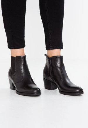 Schwarze Leder-Stiefeletten mit Blockabsätzen, getragen zu schwarzen, eng anliegenden Hosen auf weißem Hintergrund.